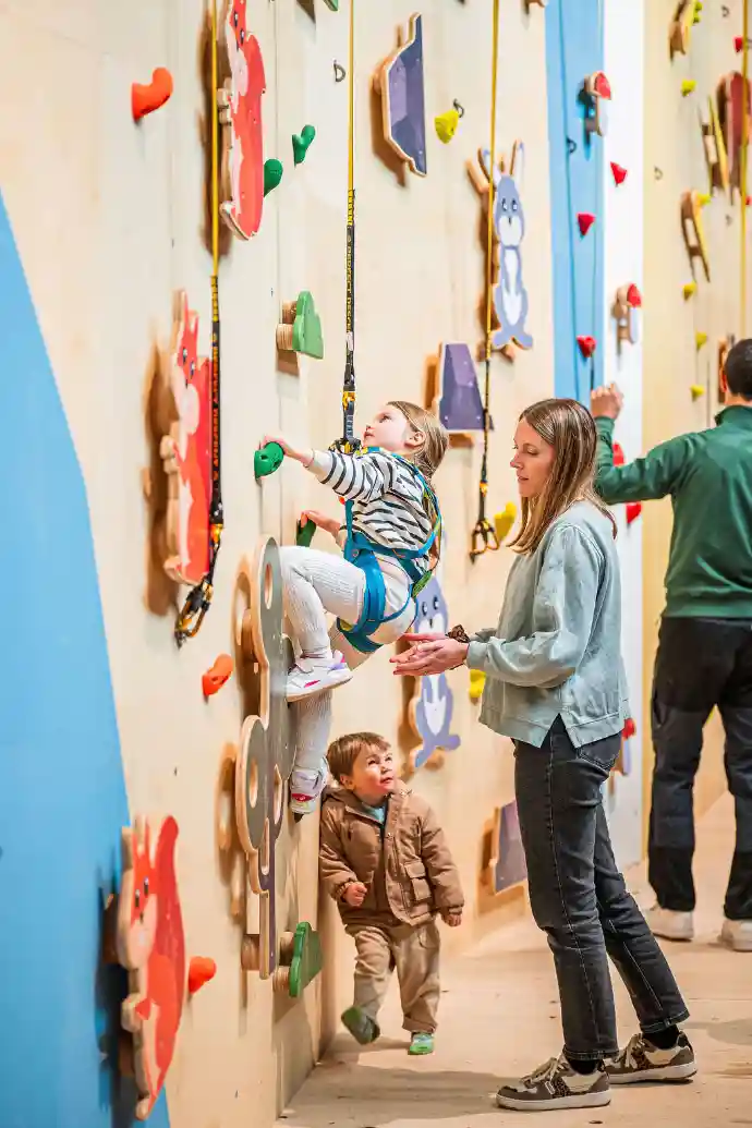 Enfant escaladant un mur avec de nombreuses prises colorées sous surveillance d'un  adulte